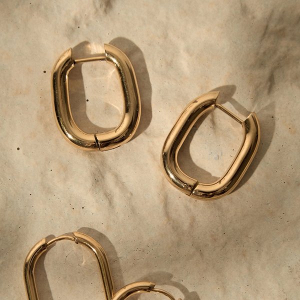 Gold-toned metal hoop earrings with a polished, rounded rectangular shape, arranged on a textured beige surface with soft shadows.