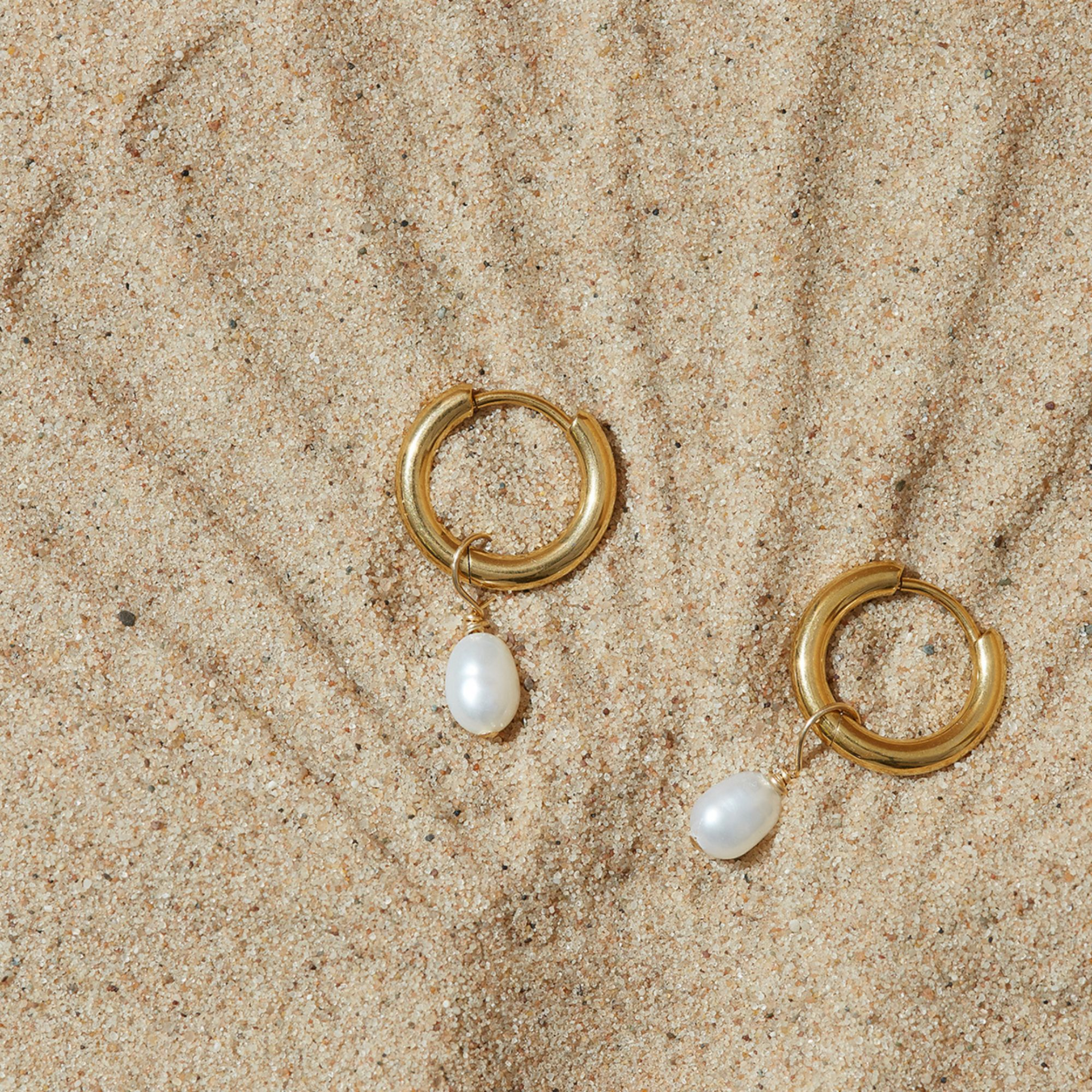 Gold hoop earrings with pearl drops on a sandy surface