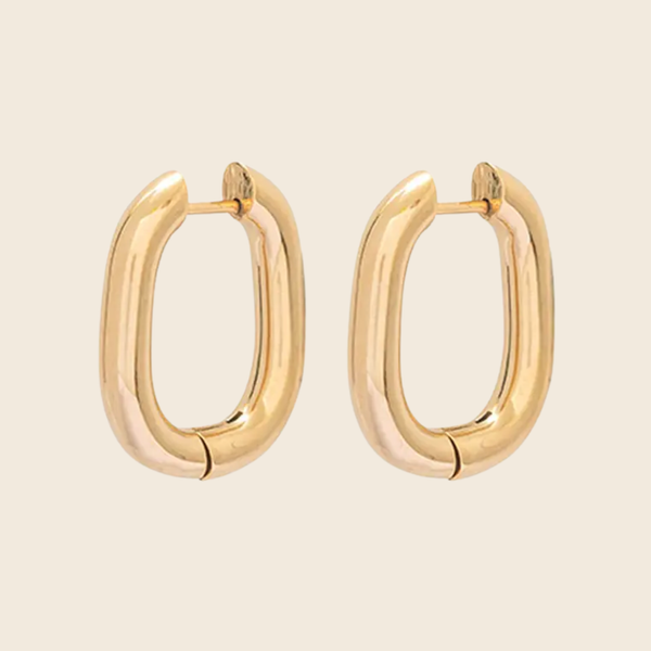 A pair of gold-toned, chunky oval hoop earrings with a polished finish, displayed on a solid light beige background.