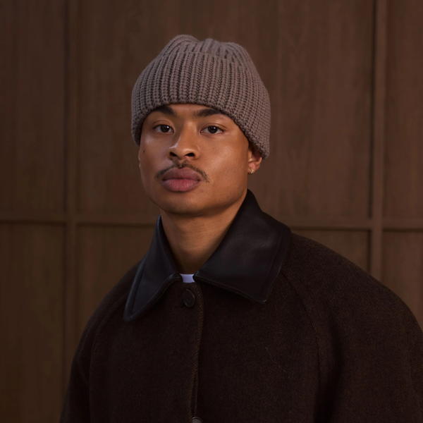 A man wearing a taupe ribbed knit beanie and a dark brown wool coat with a black collar, standing against a dark wood-paneled background.