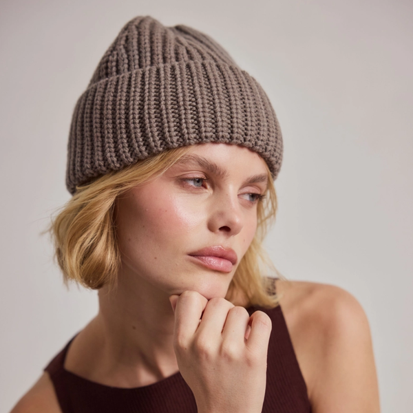 A close-up of a woman wearing a ribbed knit beanie in taupe brown, with short blonde hair and a dark brown ribbed top against a plain light grey background.