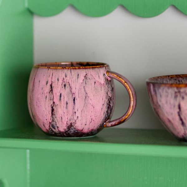 A pink ceramic mug with a brown-toned rim on a green shelf next to another matching mug.