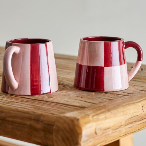 Two ceramic mugs with a glossy red and pale pink checkered pattern, displayed on a rustic wooden table against a neutral light gray background.