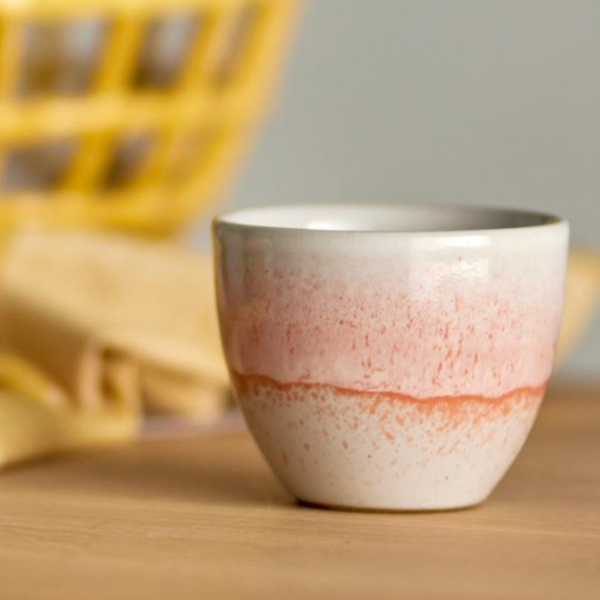 A handmade ceramic cup with a speckled pink and orange glaze over a white base, sitting on a wooden surface with a blurred yellow basket in the background.