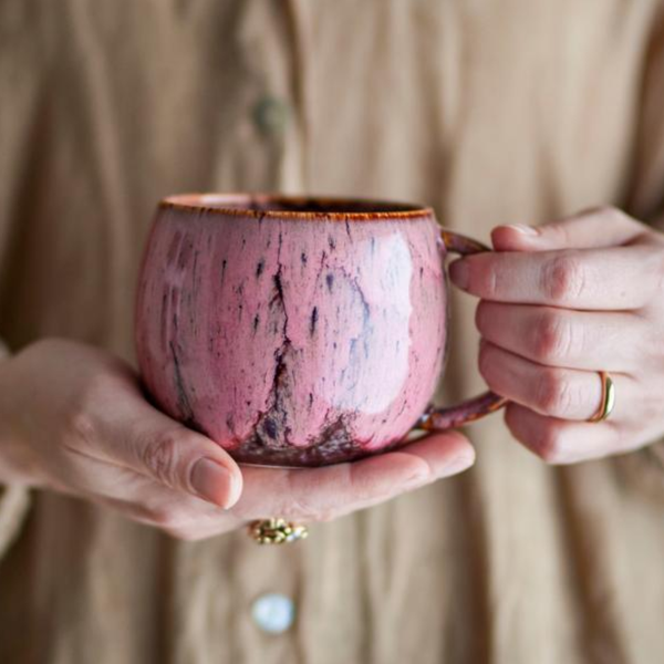 A person holding a pink marbled ceramic mug with a brown rim and handle, wearing a beige button-up shirt and gold rings.