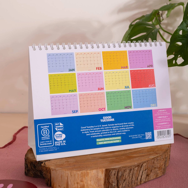 A spiral-bound desk calendar standing upright on a wooden block, showing the back side with a grid of colored months and a blue footer. The calendar is photographed indoors with a plant leaf in the background.