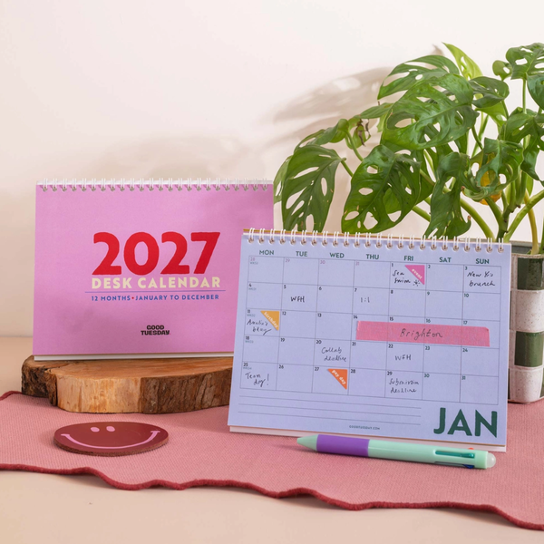 A 2027 desk calendar shown in both full monthly view and a smaller daily January spread, styled on a pink surface with a green plant and wood slice in the background.
