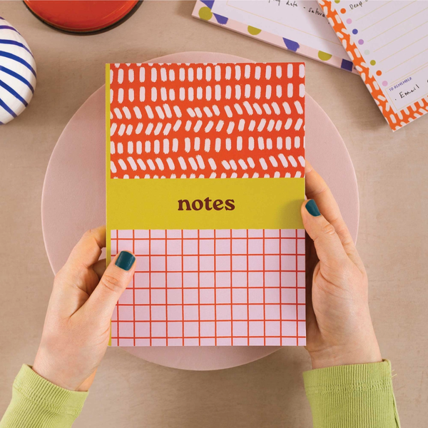 A person holds a colorful patterned notebook with the word 'notes' on the cover. The notebook has a coral-red top section with white brushstroke marks, a yellow middle band, and a pink-and-white grid bottom section.