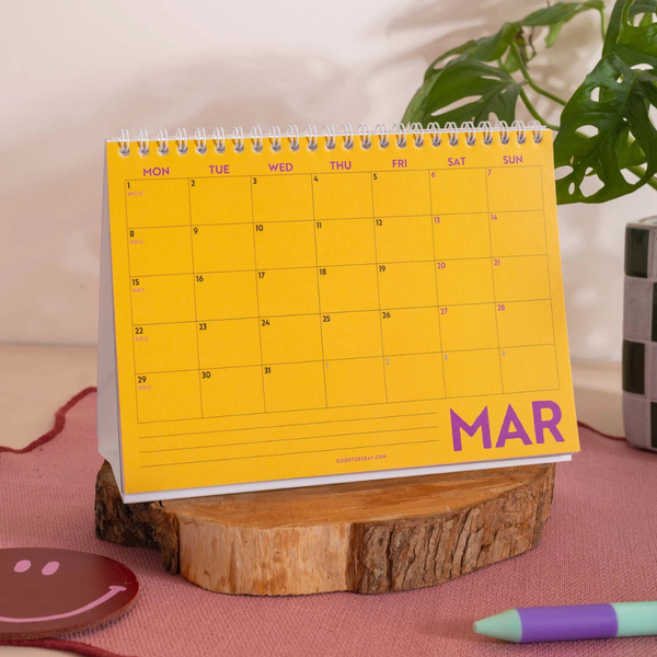 A yellow spiral-bound monthly desk calendar standing on a wooden slice, with pink lettering reading 'MAR' and a grid of days. The scene is styled on a pink surface with a plant leaf and a pen in the background.