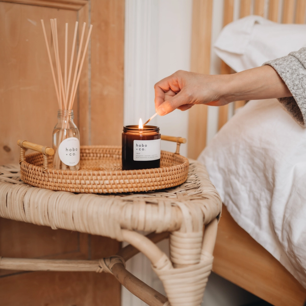 A lit amber glass candle on a woven tray beside a reed diffuser, with a person's hand lighting another candle in a cozy bedroom setting.