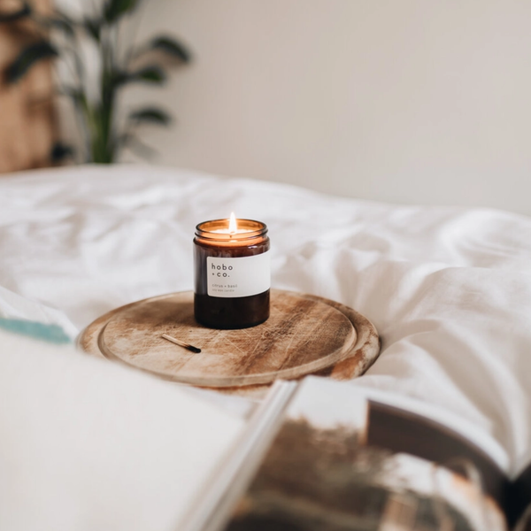 A lit amber glass jar candle with a white label reading Hobo + Co sits on a round wooden tray on a bed with white bedding and a blurred plant in the background.