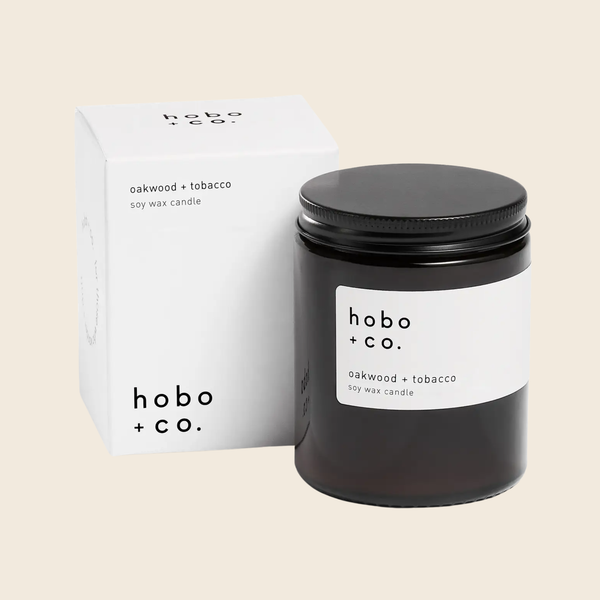 A black glass soy wax candle jar with a white label reading 'hobo + co. Oakwood + Tobacco' stands in front of its white branded box on a light beige background.