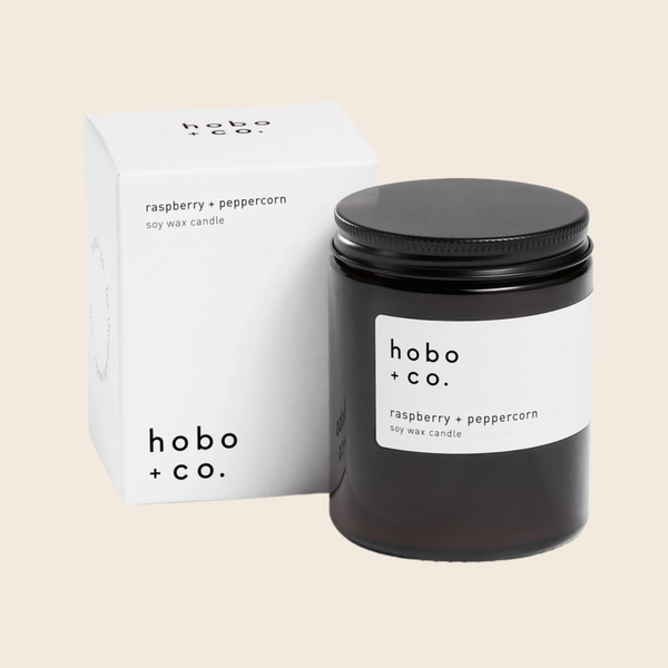 A black glass soy wax candle jar with a white label reading Hobo + co. Raspberry and Peppercorn stands in front of its white branded box on a light beige background.
