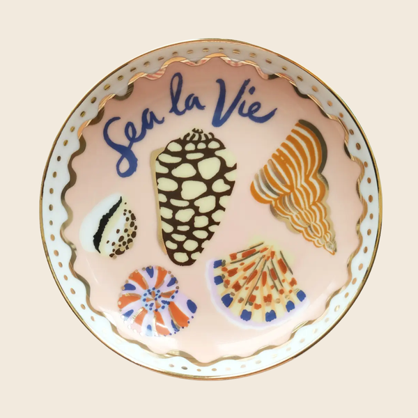 A round ceramic plate with a scalloped gold-trimmed edge and a pale pink interior, decorated with illustrated seashells and the blue script text 'Sea la Vie'.
