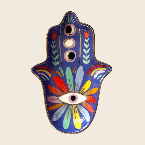 A decorative Hamsa hand tray with a vibrant hand-painted floral and eye design on a deep blue background, set against a solid light beige backdrop.