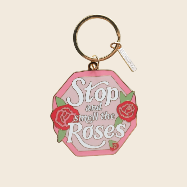 A pink octagonal keychain with the phrase 'Stop and smell the Roses' in white script, decorated with red roses and green leaves, attached to a gold key ring on a light beige background.
