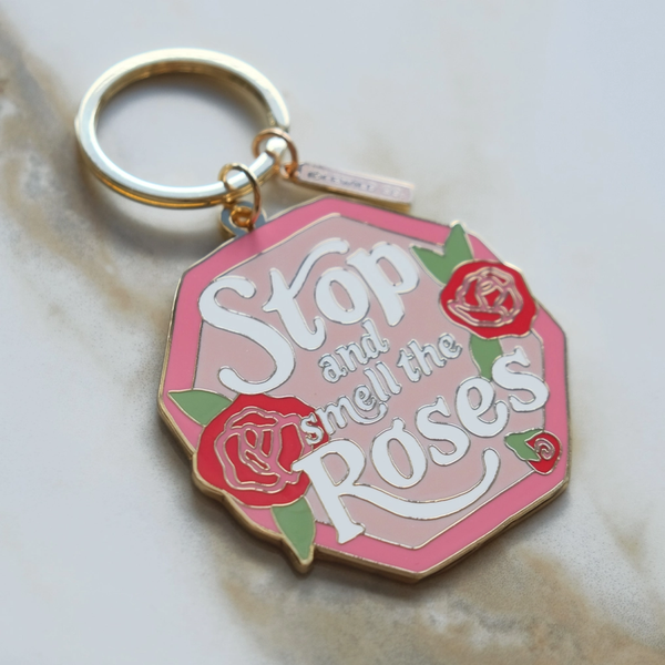 An enamel pin keychain featuring a pink and white geometric pendant with the text 'Stop and smell the roses' in white script, decorated with red roses and green leaves, attached to a gold-tone split ring.