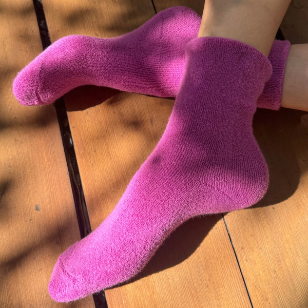 A pair of fuzzy pink socks worn on feet, resting on a wooden deck floor with dappled sunlight and shadows.