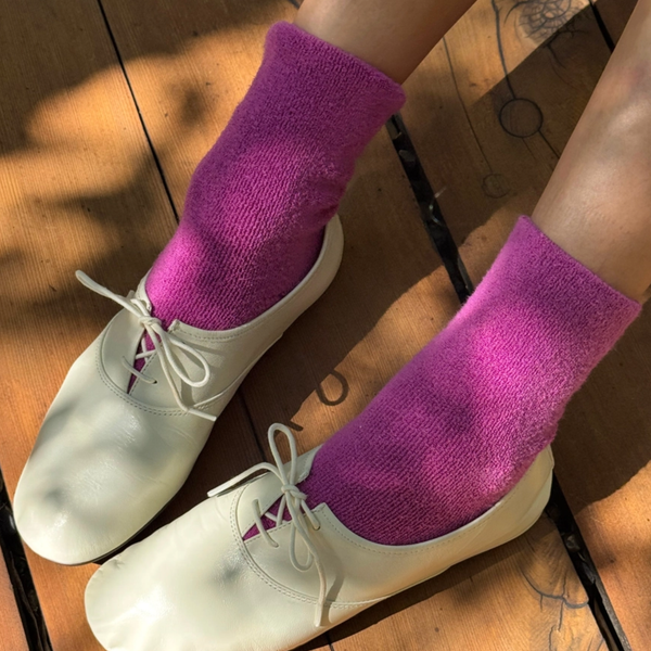A close-up of a person's feet wearing bright magenta pink fuzzy crew socks with leather lace-up shoes, standing on a wooden floor
