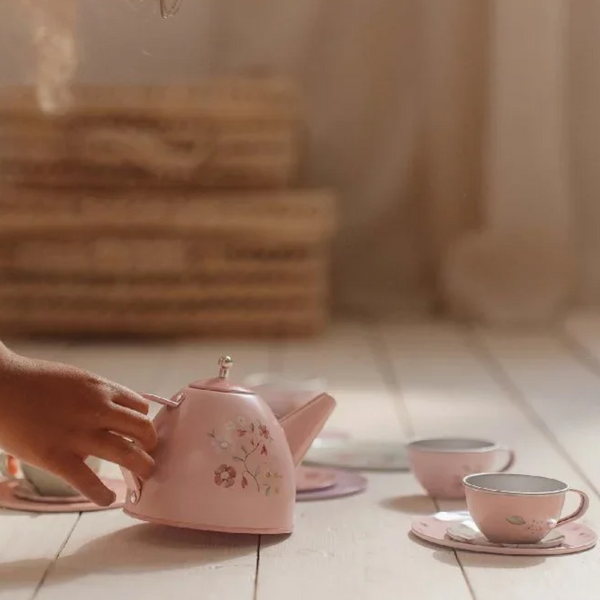 A pink metal tea kettle with a floral pattern, with a hand reaching towards it. Matching pink teacups and saucers are arranged nearby in a softly lit room.