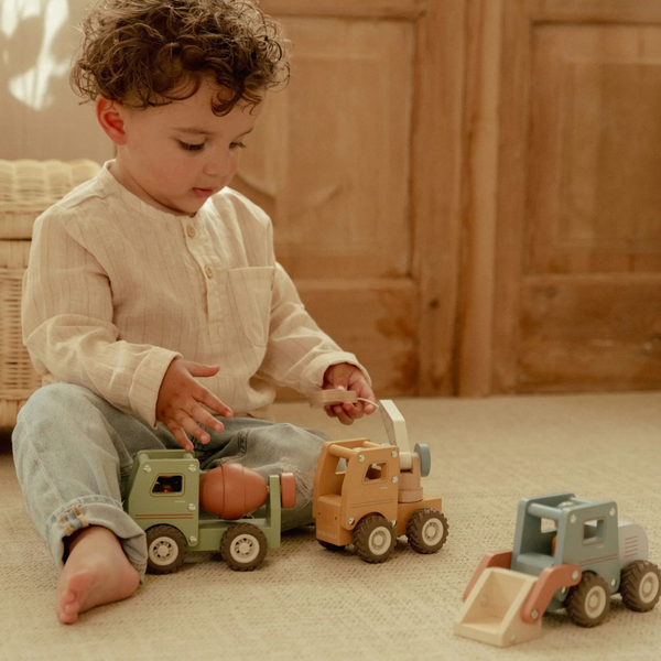 A toddler sits on a beige carpet playing with wooden toy trucks in a warm, rustic room. The child is wearing a cream ribbed long-sleeve henley shirt and light blue jeans.