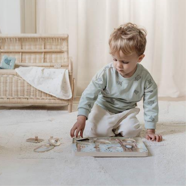 A toddler wearing a sage green long-sleeve shirt with butterfly prints and cream pants sits on a light-colored rug, interacting with a wooden toy on the floor in a softly lit nursery setting.