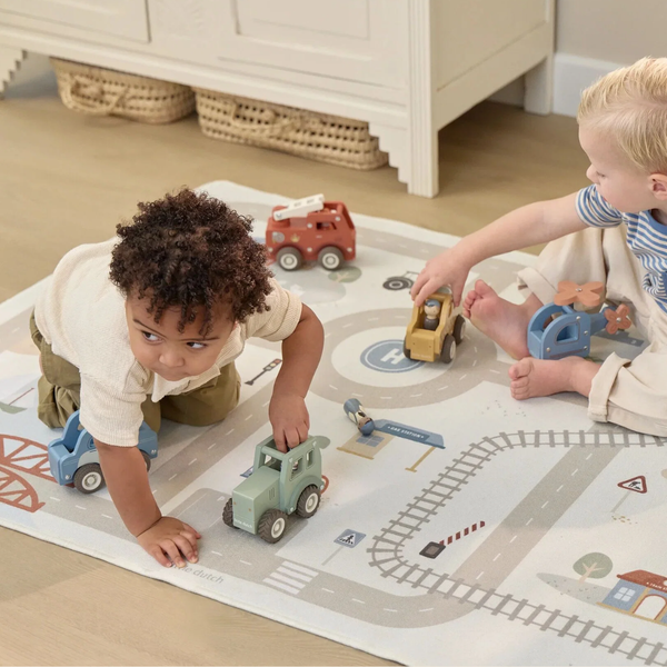 Two toddlers playing on a large road-play rug with toy vehicles in a bright indoor room. One child is kneeling and pushing a light green truck, while the other sits and reaches toward a yellow truck.
