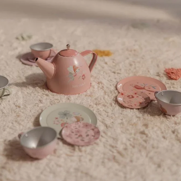 A pink metal tea set arranged on a soft cream textured rug. The set includes a teapot with a fairy illustration, cups, saucers, and plates with floral and fairy designs.