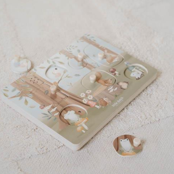 A wooden animal puzzle board with illustrated woodland scenes and small animal-shaped pieces, photographed on a soft cream fabric background.