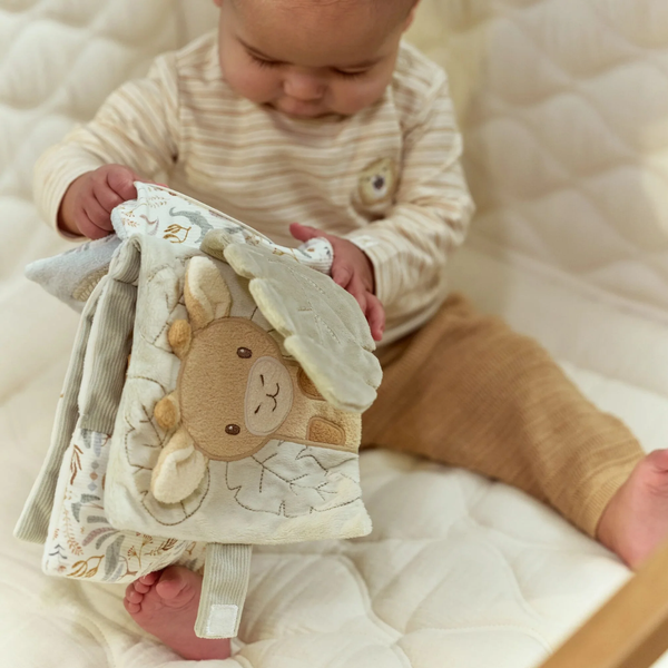 A baby sitting on a cream quilted mattress holding a soft fabric activity book with a giraffe applique.