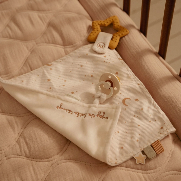 A close-up of a baby sensory blanket with a pacifier clip, featuring a cream fabric body with gold star and moon prints, embroidered text, and attached  teething toy, resting on a quilted beige mattress.