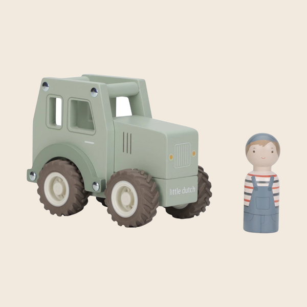 A wooden toy tractor in a muted sage green color with dark brown and cream wheels, shown next to a small wooden peg doll dressed in blue overalls and a striped shirt on a solid light beige background. The brand name 'little ducky' is printed on the side of the tractor.