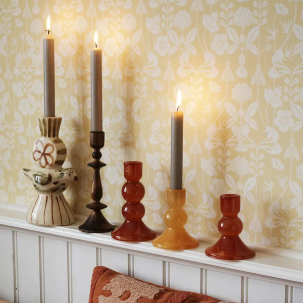 Five decorative candlesticks and taper candles arranged on a white mantelpiece against patterned wallpaper. The set includes two tall dark brown candlesticks, two amber-orange candlesticks, and one cream-and-brown patterned holder with lit candles in each.