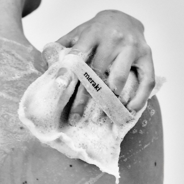 A close-up black and white photo shows a person's hand scrubbing soap onto skin, with a foam cloth carrying the visible Meraki brand name.