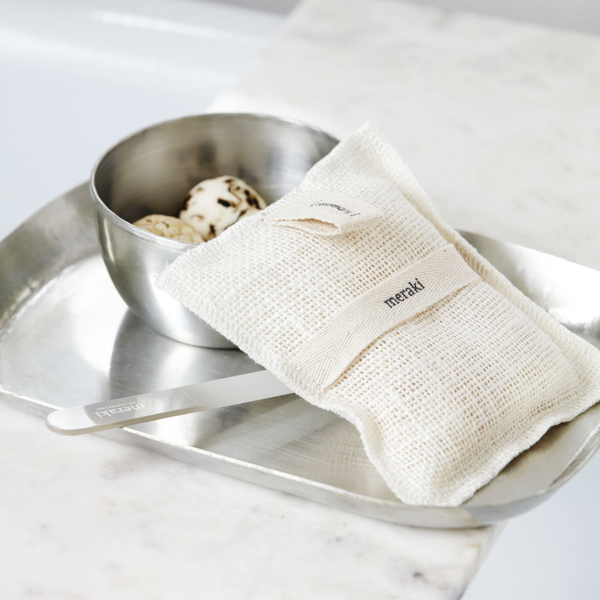 A natural beige soap bag with a fabric label reading 'meraki', resting on a silver metal tray next to a small stainless steel bowl of bath bombs on a white marble surface.