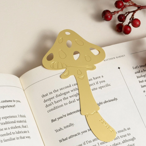 A gold metal bookmark shaped like a mushroom, clipped onto an open book with red berries in the corner as props.