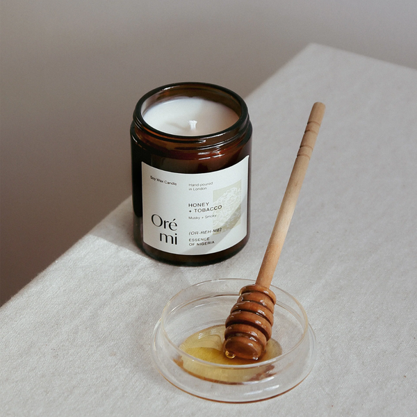Candle labeled 'Oré mi' next to a dish with honey and a wooden dipper on a light surface.
