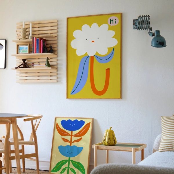 A colorful framed wall art print featuring a stylized flower with a white bloom, blue stem, and orange leaf on a yellow background, displayed in a bright living space next to a smaller matching floral print leaning on the floor.