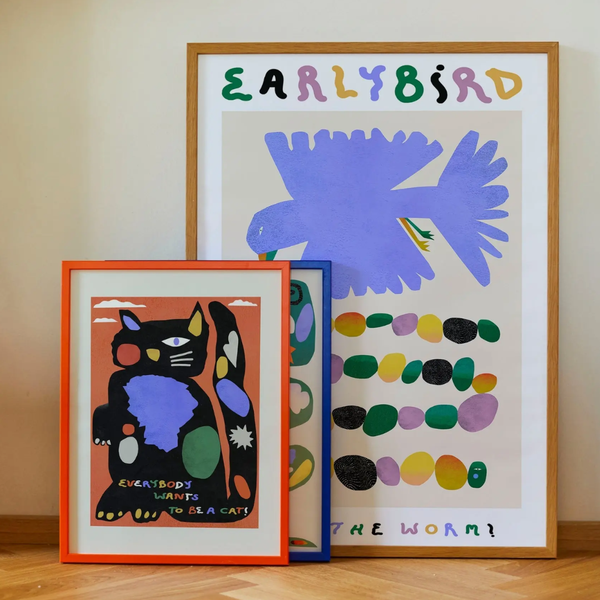 Three framed colorful art prints are arranged on a wooden floor against a beige wall. The largest print behind shows a purple bird and the words 'EARLYBIRD' and 'THE WORM!' with multicolored dots.