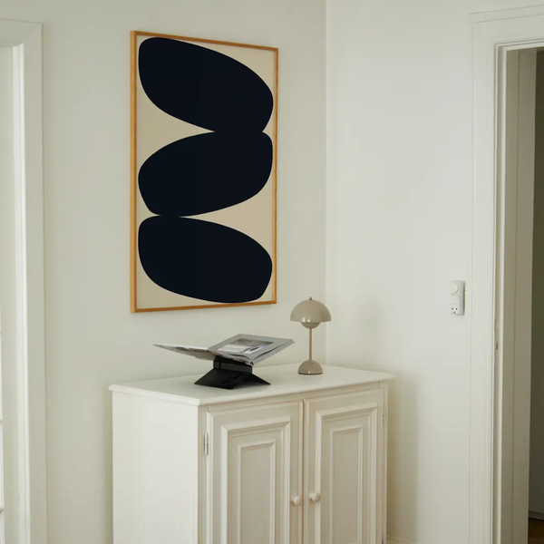 A framed abstract wall art print with three large black shapes on a warm beige background, hanging above a white cabinet in a bright, minimalist room.