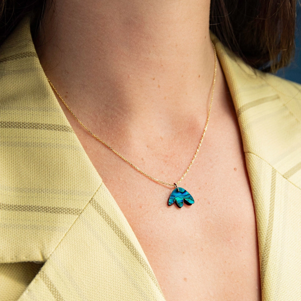 A close-up of a model wearing a delicate gold chain necklace with a small turquoise blue flower petal pendant, styled with a pale yellow striped blazer.