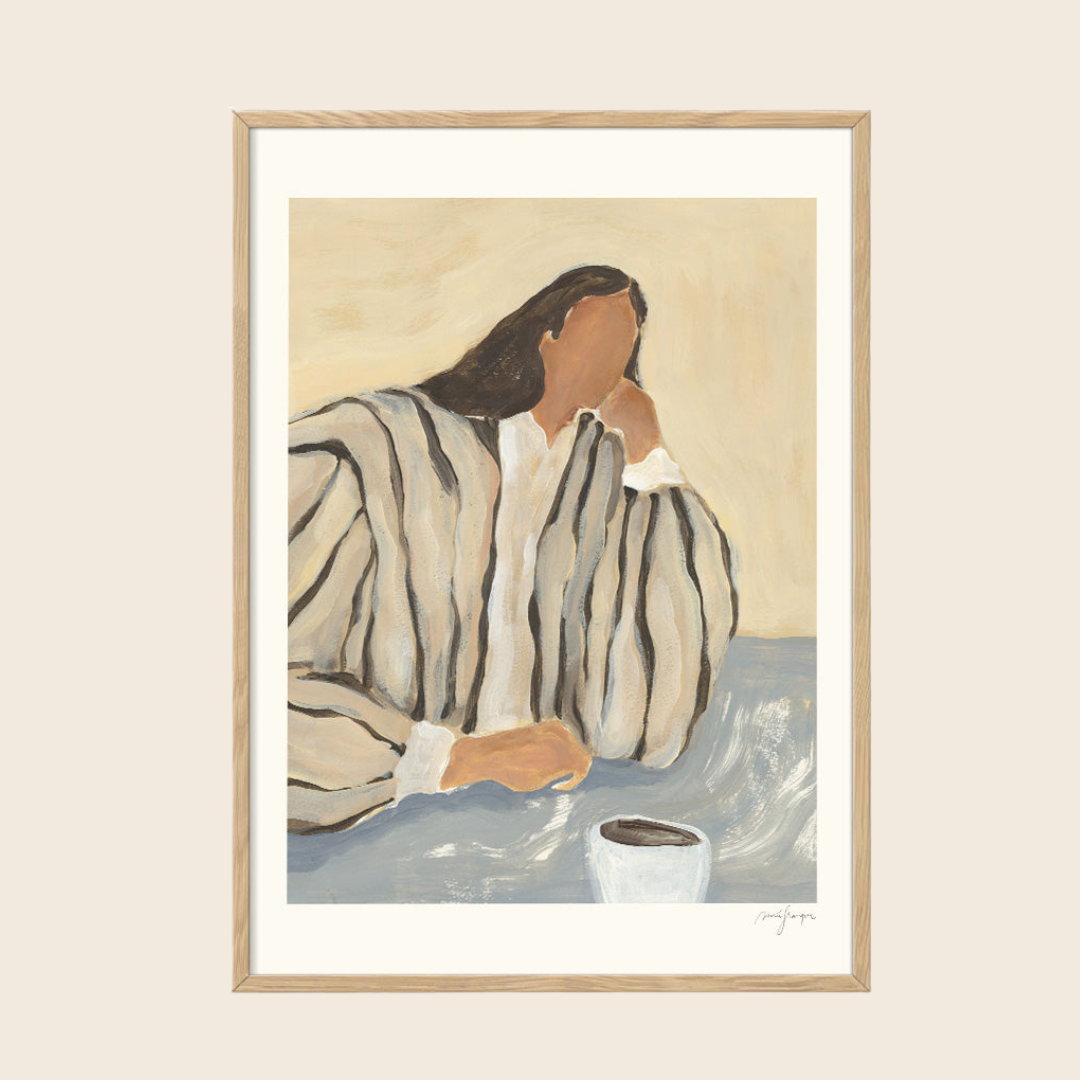 Framed artwork of a person in a robe with a cup on a table, on a beige wall.