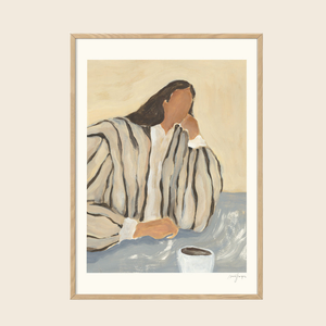 Framed artwork of a person in a robe with a cup on a table, on a beige wall.