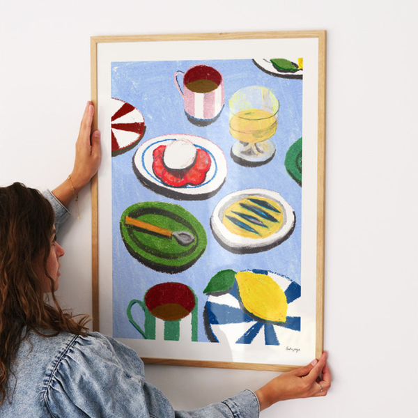 Person holding a framed colorful artwork of still life objects on a white wall