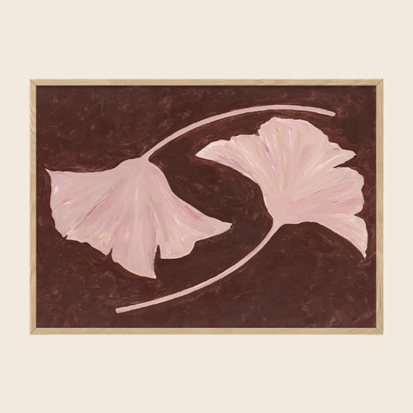 A framed art print depicting two light pink ginkgo leaves on a dark brown textured background, enclosed in a thin light wood frame against a cream-colored wall.