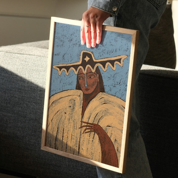 A framed art print featuring a stylised figure in a large hat against a textured blue background, held by a person in a home setting.