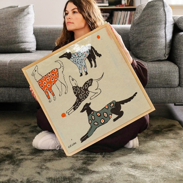 A woman sits on a gray carpet in front of a grey sofa, holding a square framed art print featuring four stylised dogs in patterned coats on a cream background.