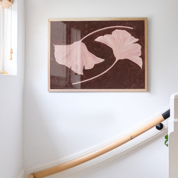 A framed art print featuring two light pink ginkgo leaves on a dark brown background, hanging on a white wall above a wooden staircase railing.