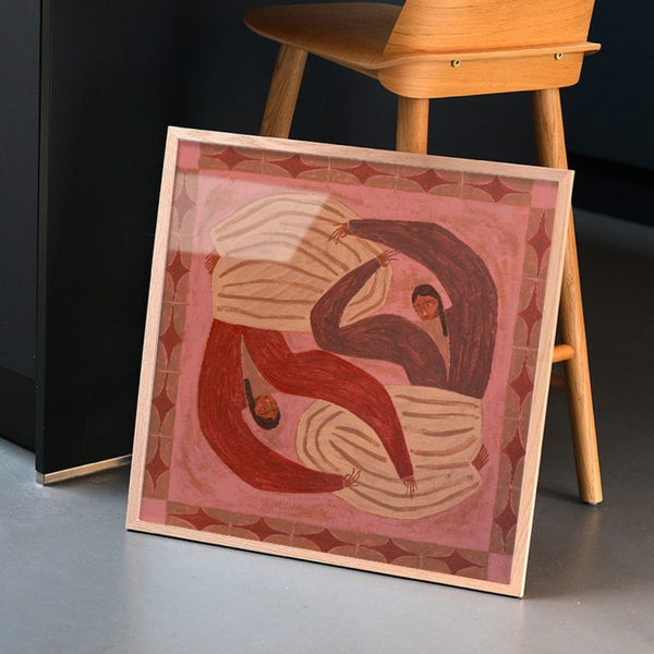 A square framed art print leaning against a black cabinet on a gray floor, with a wooden chair partially visible behind it. The artwork depicts two stylised figures in flowing red, brown, and cream garments on a pink background with a patterned border.