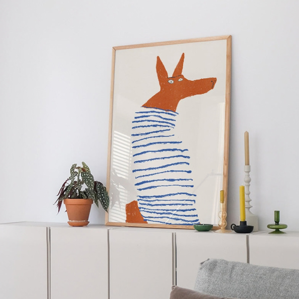 A framed art print featuring a stylised orange dog wearing a blue striped sweater, leaning against a white cabinet in a modern, minimalist room with a potted plant and candles.